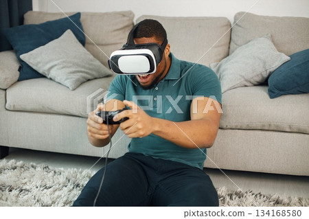 Black man in vr glasses, playing video games with virtual reality headset. Man wearing blue t-shirt. Man in virtual reality glasses sitting at home near coach. 134168580