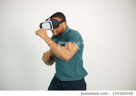 Black man in vr glasses, playing video games with virtual reality headset. Man wearing blue t-shirt. Man in virtual reality glasses standing in studio on white background. 134168589