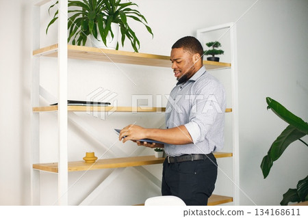 Black business man standing in office near bookshelf. Bearded man holding a folder with documents. Man wearing blue shirt. Black business man standing in office near bookshelf. Bearded man holding a folder with documents. Man wearing blue shirt. 134168611