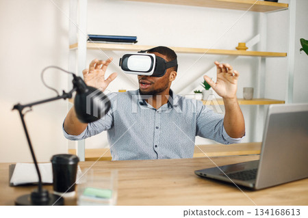 Black business man sitting in office with a laptop. Bearded man using virtual reality glases. Man wearing blue shirt. Black business man sitting in office with a laptop. Bearded man using virtual reality glases. Man wearing blue shirt. 134168613