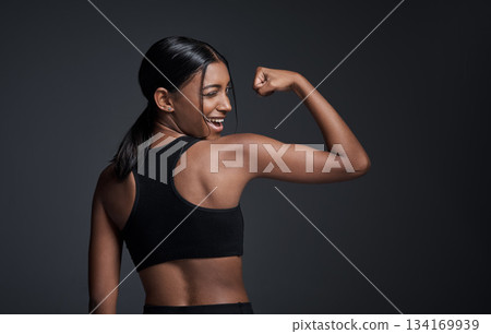 Wink, happy and woman flexing muscle in studio isolated on a black background mockup. Strong flex, funny and Indian female athlete with bicep, arm strength or bodybuilder back, fitness or workout. 134169939