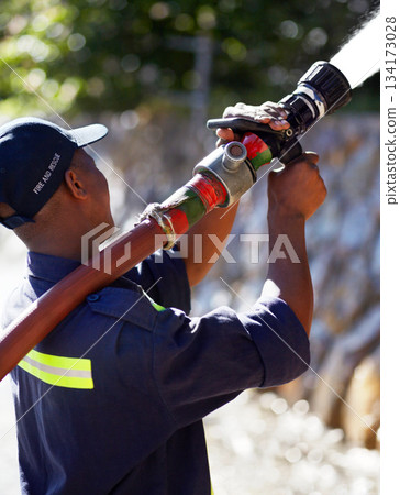 Firefighter man, water and spraying hose for emergency, rescue or firefighting services in the outdoors. Fireman using big liquid pressure pipe to spray down or put out fire outside for safety 134173028