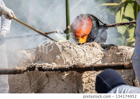Blacksmith holding a hot crucible by the iron pliers from furnace and pouring the melting gold into the statue block. 134174952