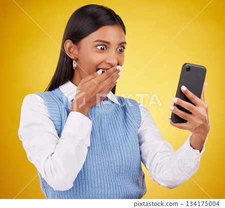 Wow, surprise and woman with phone in studio space reading text or email on yellow background. Technology, communication and good news, happy Indian girl with social media gossip or online content. 134175004