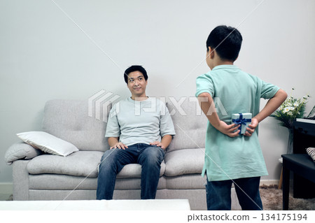 child holding gift behind their back while approaching father at home, capturing sweet surprise moment, family bonding, and warm emotional connection 134175194