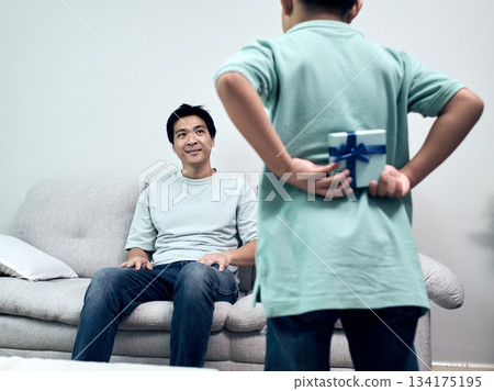 Son hiding gift behind their back while approaching father at home, portraying heartwarming surprise moment, family bonding and emotional connection 134175195