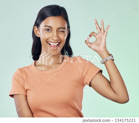 Portrait, wink and female with a perfect gesture in studio with a positive, confident and happy mindset. Happiness, smile and young Indian woman model with a ok hand sign isolated by green background Portrait, wink and female with a perfect gesture in studio with a positive, confident and happy mindset. Happiness, smile and young Indian woman model with a ok hand sign isolated by green background 134175221