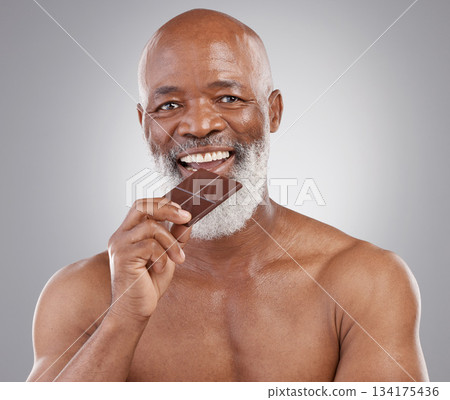 Black man, senior portrait and eating chocolate isolated on a studio background for a treat. Happy, snack smile and an elderly African model biting into a sweet candy bar for happiness and sugar Black man, senior portrait and eating chocolate isolated on a studio background for a treat. Happy, snack smile and an elderly African model biting into a sweet candy bar for happiness and sugar 134175436