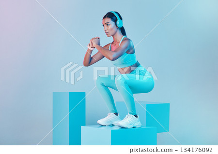 Fitness, squat and woman with focus, exercise and confident person against blue studio background. Female, model and athlete with headphones, workout goal or balance for wellness or healthy lifestyle 134176092