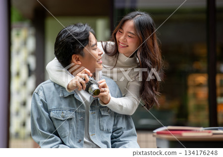 Young asian couple a woman holding camera hugging a man's neck while sitting at outdoor cafe table. Young asian couple a woman holding camera hugging a man's neck while sitting at outdoor cafe table. 134176248