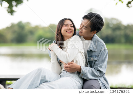Young asian couple woman getting back hug or waist holding by man sitting outdoors on bench in park. 134176261