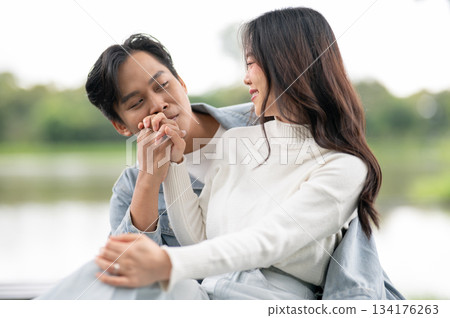 Young asian couple a man holding and kissing ring on woman's hand sitting outdoors on bench in park. 134176263