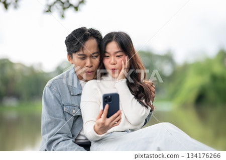 Young asian couple a man hugging woman taking selfie picture on phone while sitting outdoors in park Young asian couple a man hugging woman taking selfie picture on phone while sitting outdoors in park 134176266