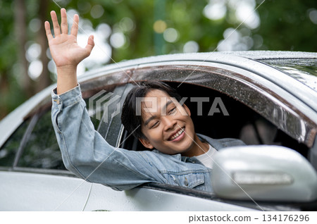 Happy young adult asian man raising arm waving a hand out of the car's window with raindrops on roof Happy young adult asian man raising arm waving a hand out of the car's window with raindrops on roof 134176296