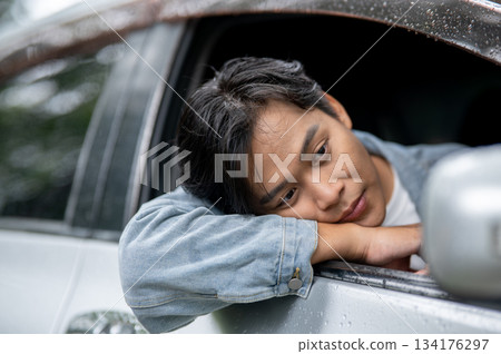 Young asian man bored waiting while resting head on arm out of the car window with raindrops on roof 134176297