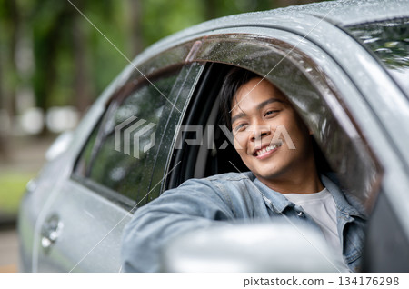 Happy young asian man looking away while putting an arm out of the car window with raindrops on roof 134176298