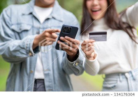 Close up young couple man pointing at phone aside woman holding credit card standing outdoor in park Close up young couple man pointing at phone aside woman holding credit card standing outdoor in park 134176304