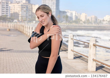 Shoulder pain, fitness woman and music of a person running by the sea with a arm injury. Workout, sports and training accident of a female runner on a exercise run and muscle strain on arms outdoor 134176487