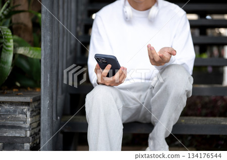 A man with explaining hand holding looking at phone while sitting outdoors at stairs of garden cafe. A man with explaining hand holding looking at phone while sitting outdoors at stairs of garden cafe. 134176544