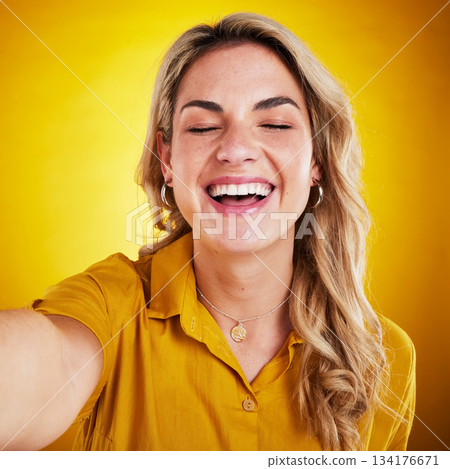 Selfie, face and smile with a laughing woman on a yellow background in studio for fun or humor. Eye closed, comic and funny with an attractive young female model enjoying comedy, being silly or goofy Selfie, face and smile with a laughing woman on a yellow background in studio for fun or humor. Eye closed, comic and funny with an attractive young female model enjoying comedy, being silly or goofy 134176671