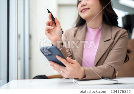 Asian business woman office worker holding pen and phone thinking over notebook sits at cafe table. Asian business woman office worker holding pen and phone thinking over notebook sits at cafe table. 134176835
