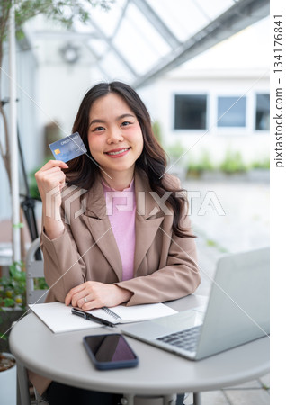 Asian business woman or office worker holding a credit card while sitting with laptop at cafe table Asian business woman or office worker holding a credit card while sitting with laptop at cafe table 134176841