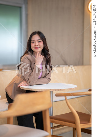 Young asian woman puts a hand under chin while sitting on sofa bench at table in cafe or coffee shop Young asian woman puts a hand under chin while sitting on sofa bench at table in cafe or coffee shop 134176874