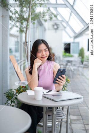 Asian woman student or freelancer holding and looking at phone aside coffee and laptop on cafe table Asian woman student or freelancer holding and looking at phone aside coffee and laptop on cafe table 134176891