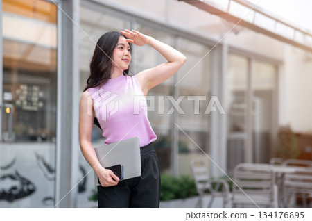 Asian woman hand covering eyes from sunlight and carrying laptop while standing in front of the cafe 134176895