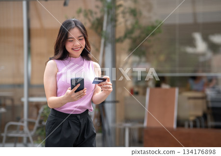 Asian young woman holding credit card looking at phone with smile while standing in front of a cafe. 134176898