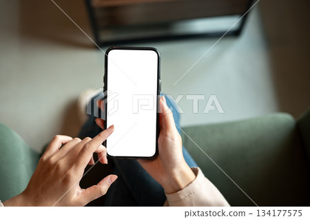 Close up of woman hand holding blank screen smartphone while sitting on green sofa in cafe or lounge Close up of woman hand holding blank screen smartphone while sitting on green sofa in cafe or lounge 134177575