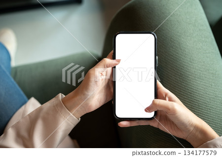 Close up woman hands holding blank screen smartphone while sitting on green sofa in a cafe or lounge Close up woman hands holding blank screen smartphone while sitting on green sofa in a cafe or lounge 134177579