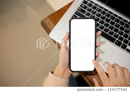 Close up woman's hand holding white screen smartphone over laptop on wooden table in cafe or library Close up woman's hand holding white screen smartphone over laptop on wooden table in cafe or library 134177580