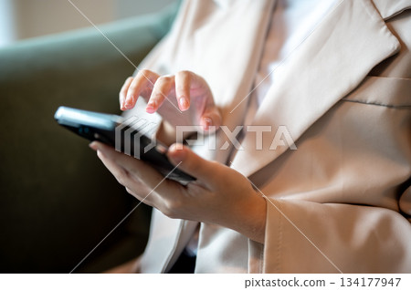 Close up of a woman holding smartphone with hand hovering over while sitting on green sofa in a cafe 134177947