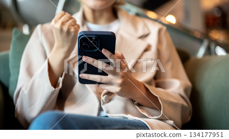 Close up of woman holding smartphone and making a fist sitting on green sofa or armchair in a cafe. 134177951
