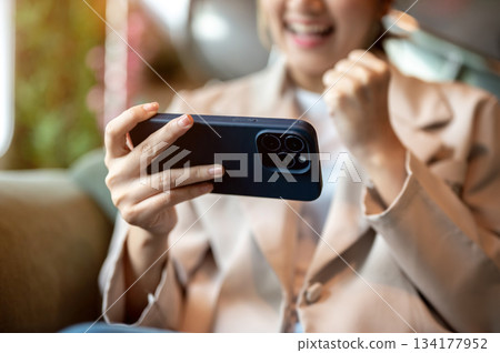 Close up of woman holding smartphone and rising a fist sitting on green sofa or armchair in a cafe. 134177952