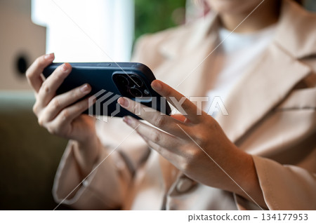 Close up of woman holding smartphone horizontally while sitting on green sofa or armchair in a cafe. 134177953