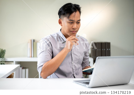 Asian businessman office worker holding pen under chin and thinking while looking at laptop on table Asian businessman office worker holding pen under chin and thinking while looking at laptop on table 134178219