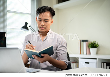 Asian businessman or office worker holding pen and looking at notebook aside laptop at working table 134178221