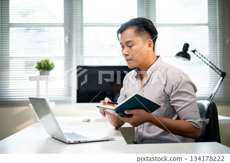 Asian businessman or office worker holding pen and notebook while looking at laptop on working table Asian businessman or office worker holding pen and notebook while looking at laptop on working table 134178222