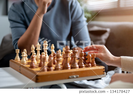Close up of woman's hand choosing a chess piece on wooden chessboard playing with man at sofa table Close up of woman's hand choosing a chess piece on wooden chessboard playing with man at sofa table 134178427