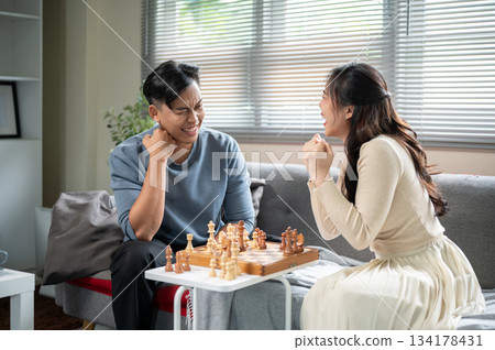 Asian woman laughing after winning a chess match on wooden board playing with a man at sofa table. Asian woman laughing after winning a chess match on wooden board playing with a man at sofa table. 134178431