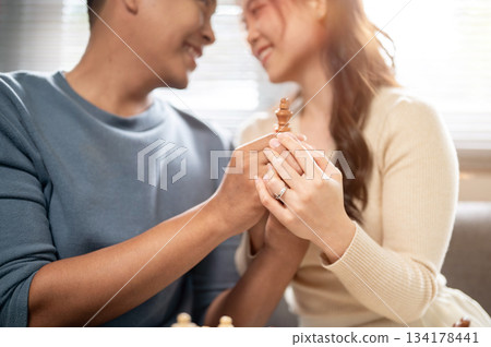Close up smiling asian man and woman holding a wooden chess piece together as sitting on home's sofa 134178441