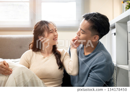Asian woman looking and poking man's cheek while sitting together on floor aside sofa in living room Asian woman looking and poking man's cheek while sitting together on floor aside sofa in living room 134178450