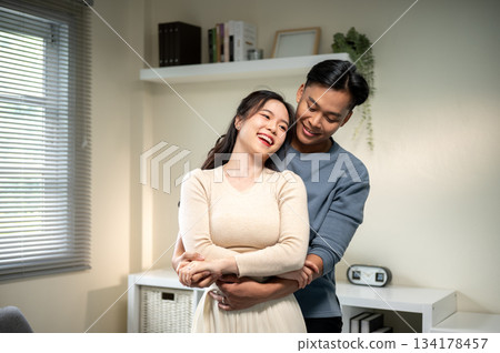 Sweet couple smiling asian man and a woman holding hands and standing hugging in living room at home Sweet couple smiling asian man and a woman holding hands and standing hugging in living room at home 134178457