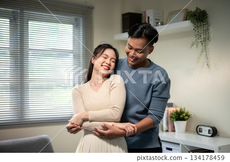 Sweet couple smiling asian man and woman holding hands while standing hugging in living room at home Sweet couple smiling asian man and woman holding hands while standing hugging in living room at home 134178459