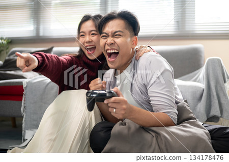 Asian man holding console making a fist in excite after winning game with woman hugging his shoulder Asian man holding console making a fist in excite after winning game with woman hugging his shoulder 134178476