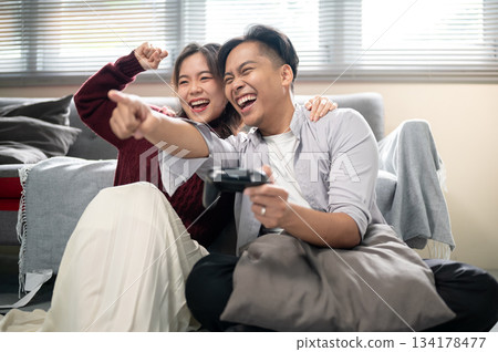 Asian man holding console playing game and pointing aside a cheering woman on a living room's floor. 134178477