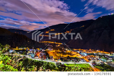Illuminated winding road of Via a Runtun rises above Banos de Agua Santa at night in Ecuador. Scenic landscape features glowing city lights and dark Andean mountains 134178591