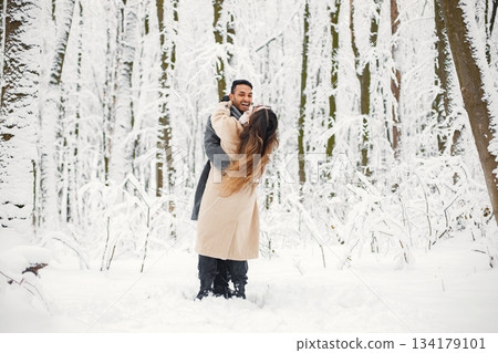 Indian man and caucasian woman walking in winter forest together. Lovely couple dancing on a snow. Man wearing grey coat and woman beige coat. Indian man and caucasian woman walking in winter forest together. Lovely couple dancing on a snow. Man wearing grey coat and woman beige coat. 134179101